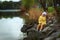 Small 5 years old girl in yellow coat and white hat sits on the river bank in spring time. Child on rock near water. Thinking
