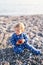 Small baby in a blue overalls sits on a pebble beach and holds an apple. Close-up