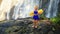 Small Blond Girl Stands on Stone against Large Waterfall