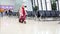 Small blonde girl in a pink winter coat with red suitcase walking across waiting hall in the airport.