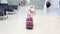 Small blonde girl in a pink winter coat with red suitcase walking across waiting hall in the airport.