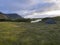 Small blue tent standing alone on green grass on mossed creek banks in Hornstrandir Iceland, snow patched hills and cliffs,