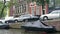 Small boats with covers moored at the side of a canal with cars parked in the