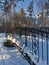 A small bridge with forged cast-iron trellises over a frozen river on a sunny winter day against the backdrop of snow-covered