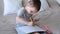 A small caucasian boy in a gray T-shirt and shorts on the bed draws with colored pencils in a sketchbook.