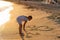 Small Caucasian girl drawing figures of hearts with a stick on sandy beach at summer sunset