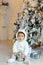 Small charming baby boy clothing polar bear cub sits near a Christmas fir-tree, holding a ball in the interior of the house