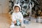 Small charming baby boy clothing polar bear cub sits near a Christmas fir-tree, holding a ball in the interior of the house