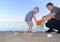 A small child collects trash on the beach. His dad points his finger where to throw garbage. Parents teach children cleanliness.