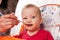 A small child eats with a spoon and sits and holds the edge of the feeding table in the nursery, grimy and stained with food, baby