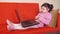 Small child playing on the laptop on an orange couch at home. The little girl is engaged on a large metal laptop on the stand for.