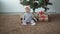 A small child in a Santa Claus hat sits on the floor near the Christmas tree. Lots of boxes with gifts around him