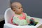 A small child sits on a white chair with a toy in his hands, against the background of a gray wall. Profile of a serious little