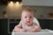 A small child in a white bodysuit sits in a high chair in the kitchen and waits to eat