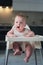 A small child in a white bodysuit sits in a high chair and yawns