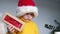 A small child in a yellow sweater and a red Santa Claus hat holds a gift in his hand and lifts it up.