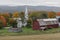 A small church sits on a farm next to a weathered red barn during Autumn