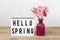 Small colored pink carnations in vase, lightbox with text Hello Spring, flamingo figure on wooden table and grey wall background