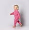 Small cute blonde smiling cheerful baby girl in pink warm comfortable jumpsuit barefoot walking over grey wall background