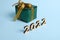 Small gift box wrapped in shiny glitter green wrapping paper and gold ribbon and bow on blue background with wooden numbers year