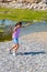 Small girl in pastel clothes running along the riverbank with a reed in hand