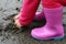 Small girl in pink rubber boots grabs wet mud from puddle
