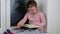 Small girl sits on windowsill behind the frozen window. Cute little girl sits on window ledge and reads the diary.