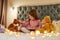 A small girl wearing pyjamas sitting alone on a huge bed cross-legged barefoot holding a teddybear`s ear with her