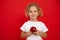 Small little expressive, positive curly blonde girl holding red juice apple in hands looking at camera on red backview