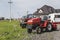 A small mini red tractor stands on a farm yard on green grass and waits for work to begin against another tractors and