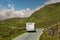 Small motorhome on a small narrow asphalt road in a mountains. Wool sheep grazing grass on the road side. Warm sunny day, cloudy