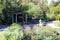 A small pergola with a brown top near a circular water fountain surrounded by lush green plants and trees in the garden