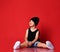 Small positive boy in stylish casual clothing, cap and white sneakers sitting on floor, looking up and smiling over red wall