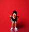 Small positive boy in stylish casual clothing, cap and white sneakers standing crouching over red wall background