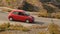 Small red car quickly appearing and driving down road behind hills, travel
