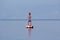 Small red navigational buoy with light on top and solar panels for charging surrounded with calm blue sea