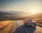 A small red retro car drives off into the distance towards the hills above which there is a huge orange sun, tourist suitcases on