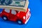 A small red toy car carries on the roof a stack of paper banknotes, blue background. The concept of costs for maintenance, car