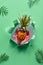 Small ripe orange pineapple cradled in human hands. Hands with the fruit show out of torn paper hole. Tropical green geometric