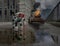Small robot standing in puddle with burning tank in the background