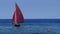 Small sailboat, an old sailing dinghy with dark red sails floats in the blue sea