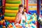 Small smiling girl in dress sitting in colorful soft decorative balls in playroom