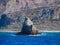 Small stony island - waves crashing - big rocky cliffs in the background - deep blue sea