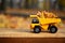 A small toy yellow truck is loaded with yellow fallen leaves. The car stands on a wooden surface against a background of a blurry