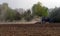 A small tractor cultivates a plowed field in the forest. Dust trail behind the tractor.