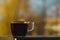 Small transparent cup of black coffee. Placed on dark wooden table in front of window. Bright blurred autumn sky and yellow trees