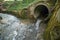 Small water stream running out of an industrial-size concrete pipe in a forest in Europe. Daytime, no people, wide angle