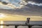 Small white brown pet dog in pink jacket seated on the bench on rocky mediterranean sea shore at sunrise with epic cloudy sky