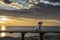 Small white brown pet dog in pink jacket seated on the bench on rocky mediterranean sea shore at sunrise with epic cloudy sky