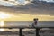 Small white brown pet dog in pink jacket seated on the bench on rocky mediterranean sea shore at sunrise with epic cloudy sky